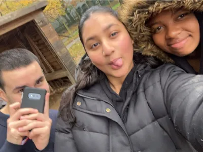 Selfie of three people; a man in the foreground taking the photo with a phone, a lady in the centre sticking her tongue out, and another lady on the right smiling under a fur-trimmed hood. They are outdoors with a wooden structure and trees with yellow leaves in the background.
